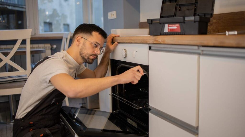 Man in overalls repairing a built-in oven with a screwdriver in a modern kitchen